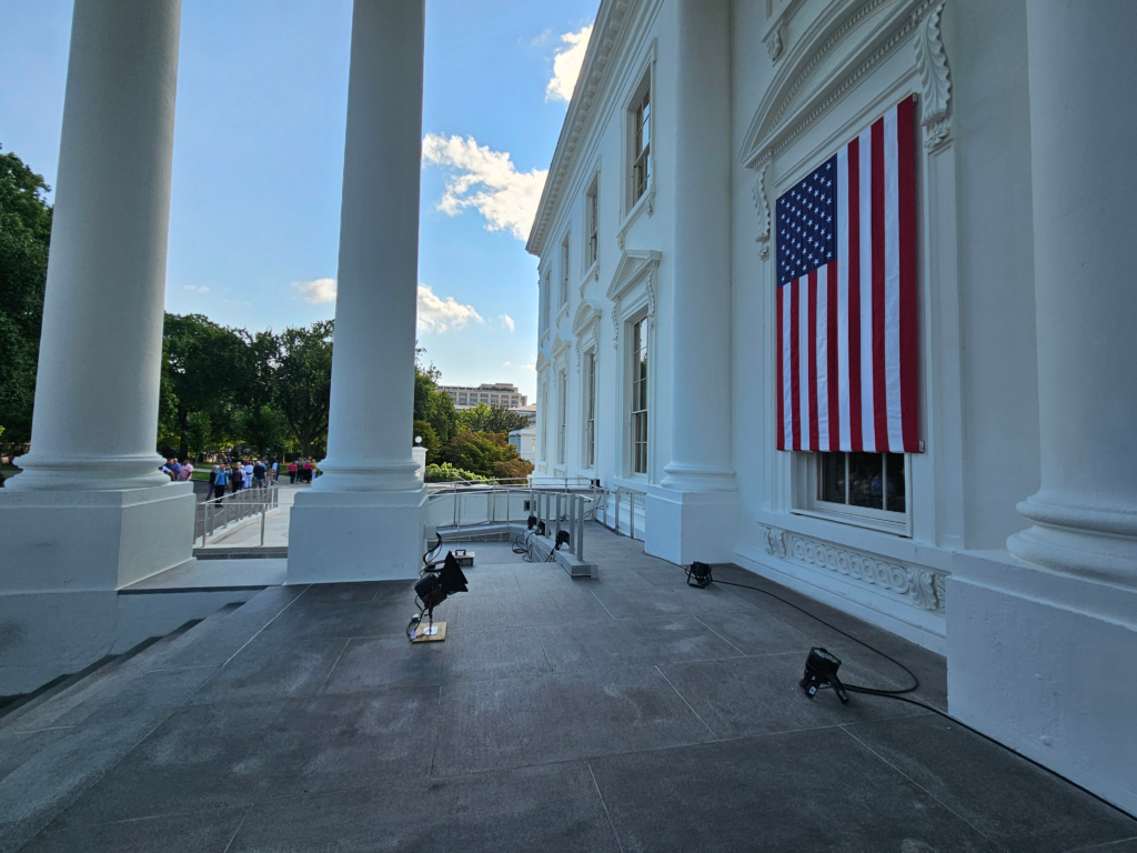 Exterior of White House with a close-up of the American Flag hanging vertically and columns on the side.