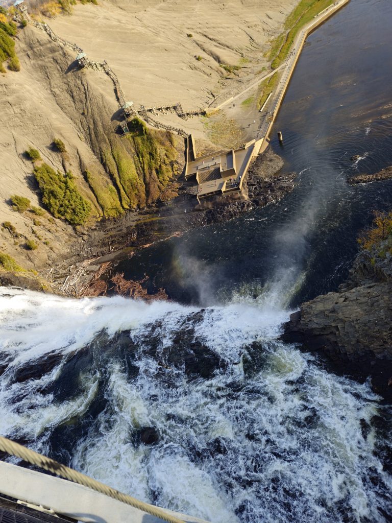 A view of Montmorency Waterfall as the waters crush into the St. Lawrence River below. A sandy mountain with wooden steps climbing to the top can be seen in the distance.